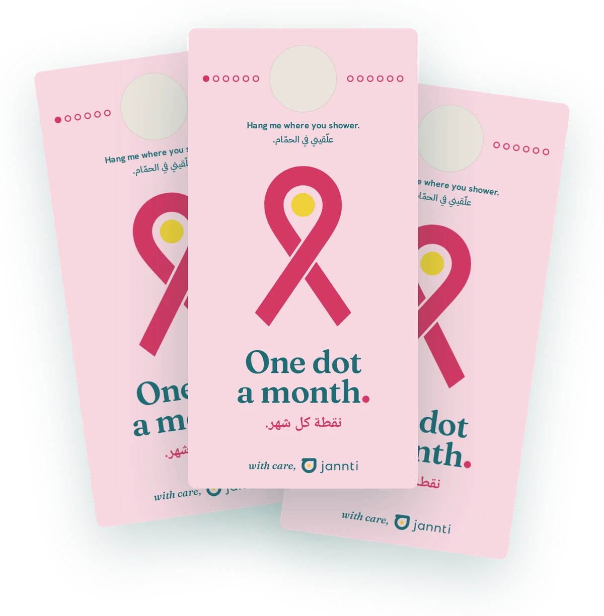 Three One Dot a Month shower cards, fanned out. Each is blush pink with a hanging hole and a row of twelve monthly dots at the top, Hang me where you shower in English and Arabic, the pink ribbon holding the yellow dot, One dot a month in English and Arabic, and with care, jannti.